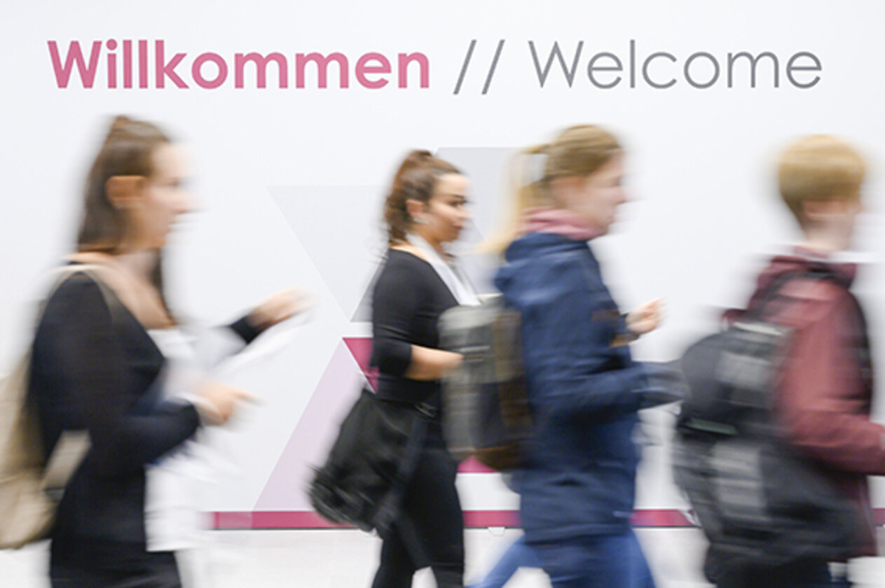 Menschen auf dem Weg in die Messehallen zur expopharm. Darüber ist der Schriftzug Willkommen/Welcome zu lesen.