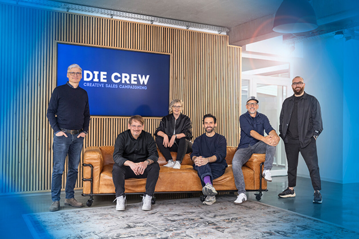 Eine Frau und drei Männer sitzen auf einem Sofa, zwei Männer stehen neben dem Sofa.