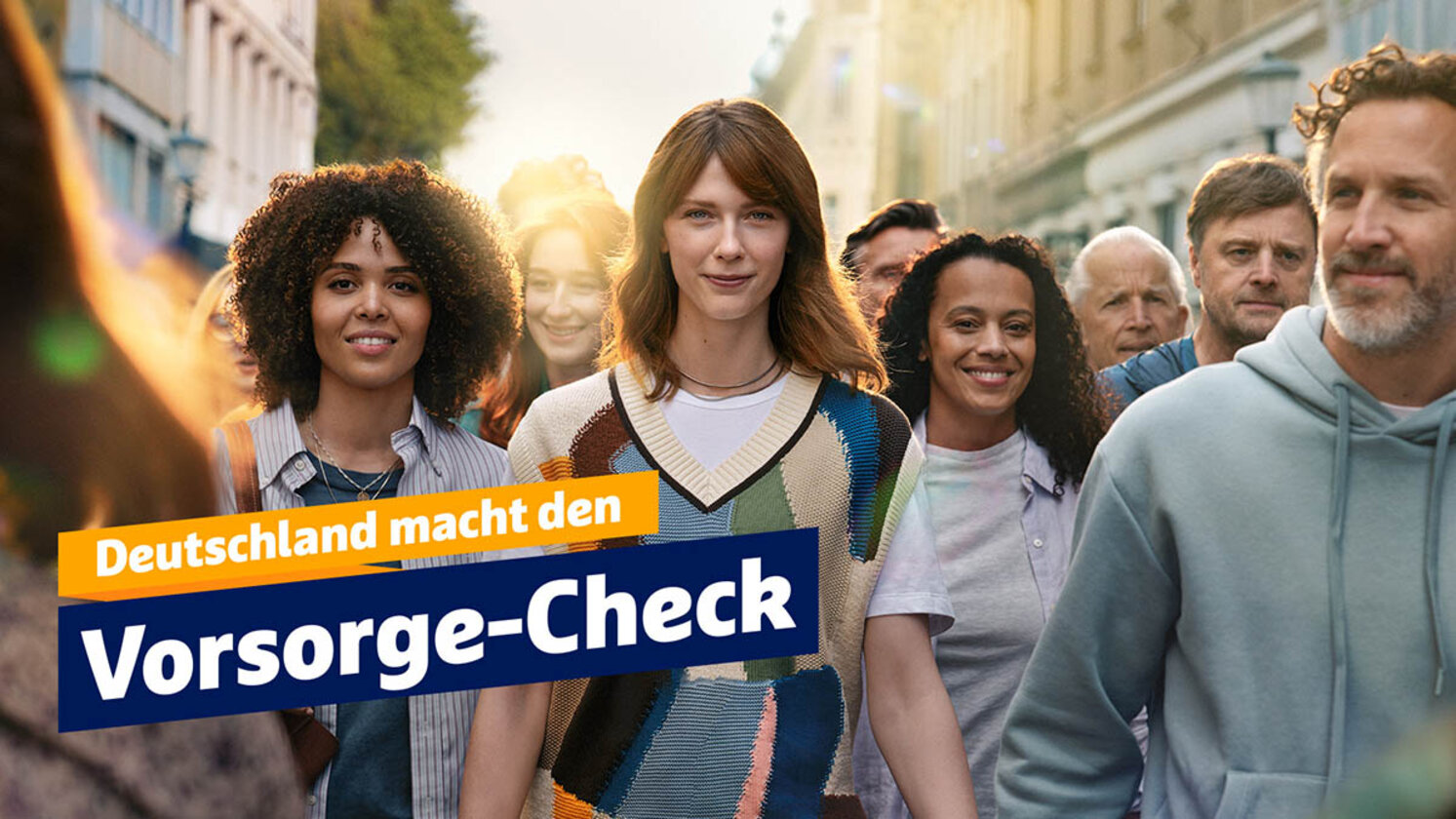 Das Bild zeigt eine Gruppe von Menschen unterschiedlichen Alters und ethnischer Herkunft, die gemeinsam auf einer sonnigen Straße spazieren. Im Vordergrund sind drei Frauen, eine mit lockigem Haar und dunklem Teint, eine mit glattem, rötlichem Haar und heller Haut, und eine mit lockigem Haar und mittlerem Teint, die alle lächeln. Im Hintergrund sind weitere Menschen zu sehen, die ebenfalls lächeln und in Richtung Kamera blicken. Auf der linken Seite des Fotos steht der Text "Deutschland macht den Vorsorge-Check" in weißen, gefetteten Buchstaben auf einem blau-gelben Banner.