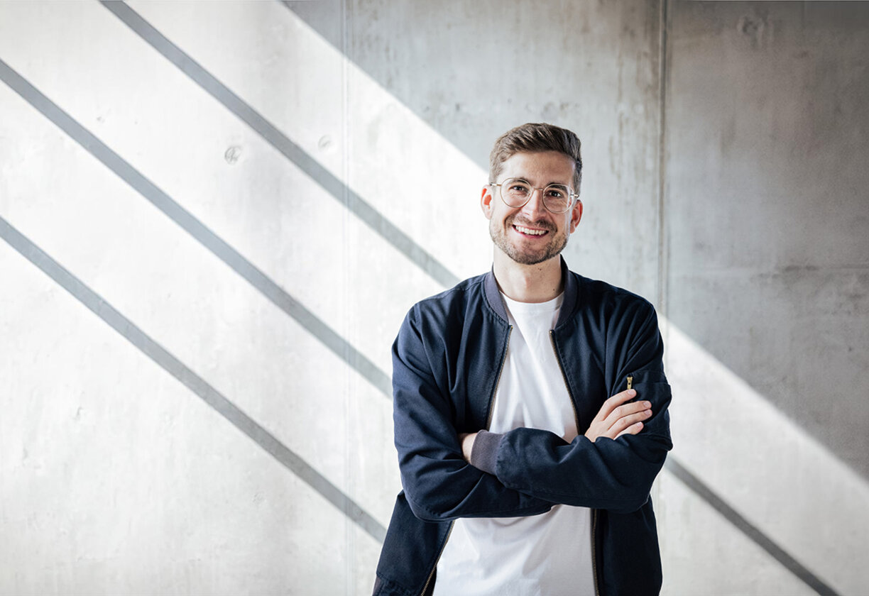 Ein brilletragender Mann steht mit verschränkten Armen vor einer sonnenbeleuchteten Betonwand. Er trägt ein weißes T-Shirt mit einer dunkelblauen Jacke darüber und lacht offen.
