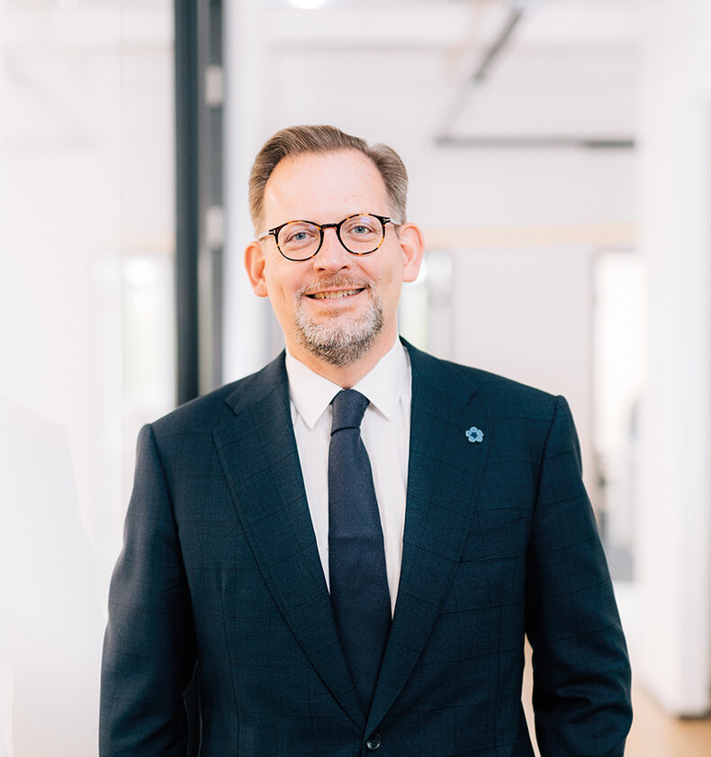 Ein lächelnder Mann mittleren Alters in einem dunklen Anzug und einer Krawatte steht in einem modernen Büro. Er trägt eine Brille und hat kurze, leicht graue Haare sowie einen gepflegten Bart. Im Hintergrund ist unscharf ein heller, freundlicher Büroraum zu erkennen.