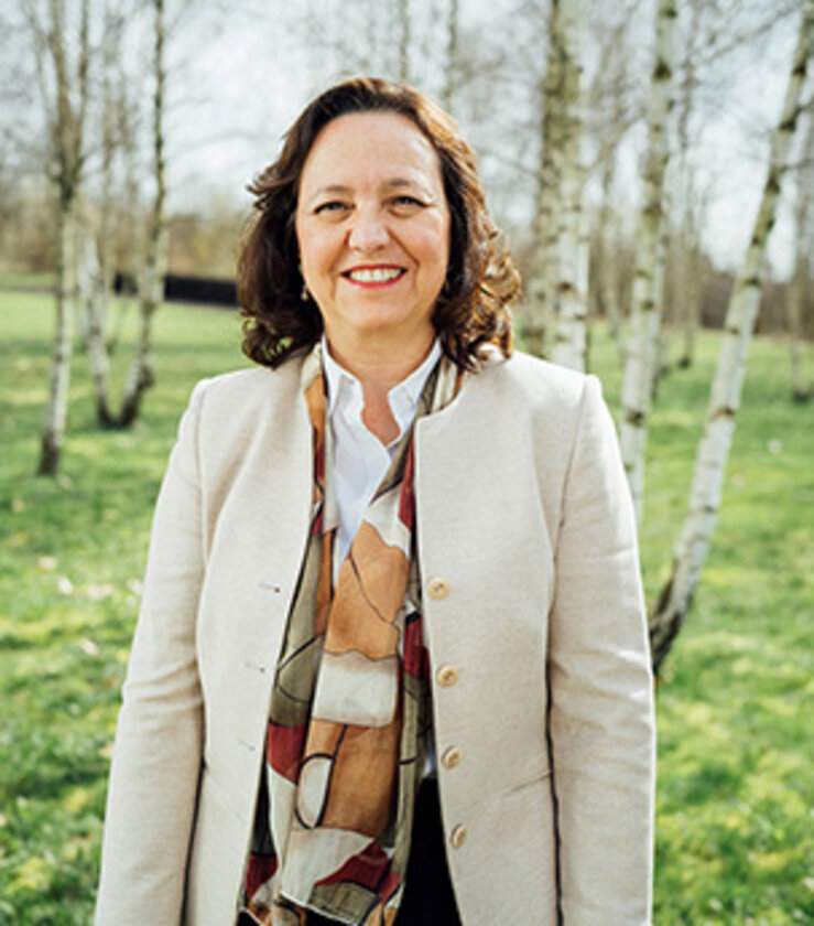 Mittelalte Frau mit dunklen, schulterlangen Haaren steht zwischen Birken. Sie trägt eine Bluse, darüber einen hellen Blazer. Sie lacht.