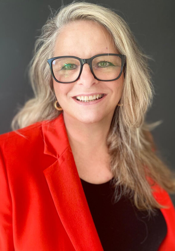 Frau mit blonden, langen Haaren, schaut lachend in die Kamera. Sie trägt einen roten Blazer und eine schwarze Brille