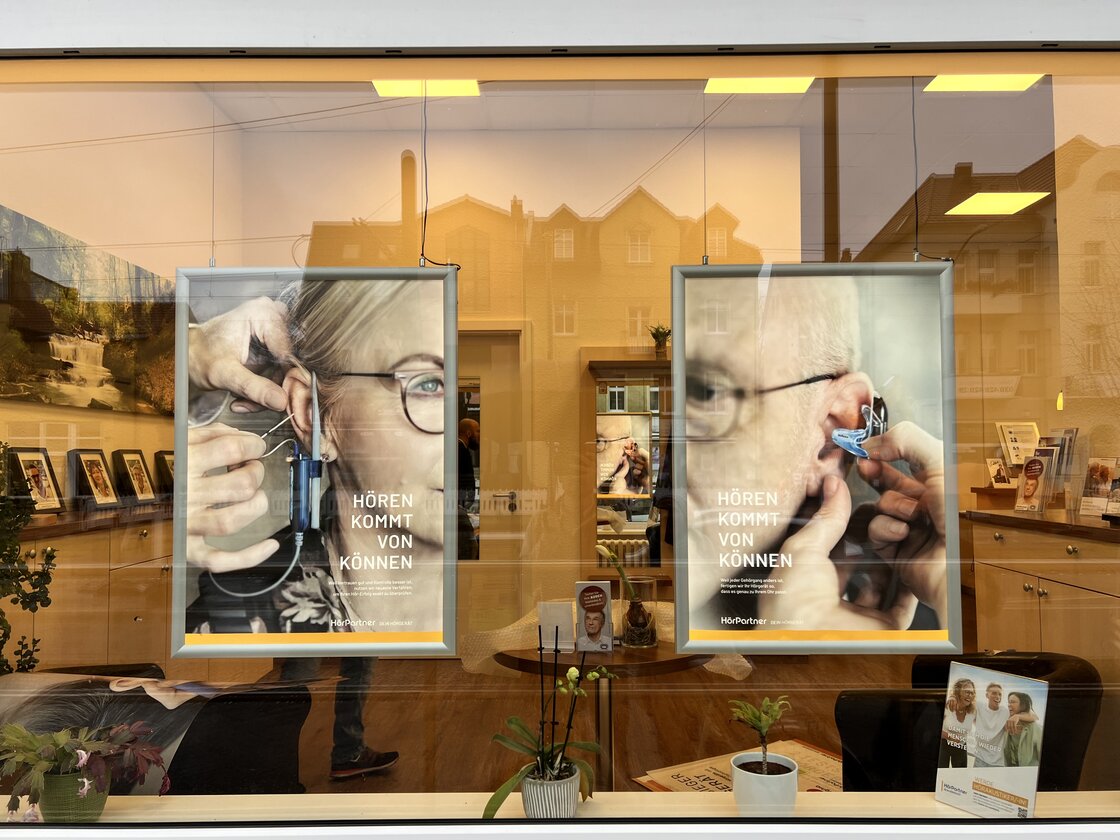 In einem Schaufenster hängen 2 Kampagnenbilder mit Menschen mit Hörgerät.