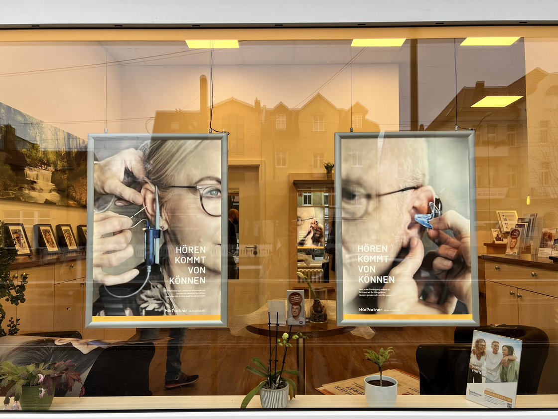 In einem Schaufenster hängen 2 Kampagnenbilder mit Menschen mit Hörgerät.