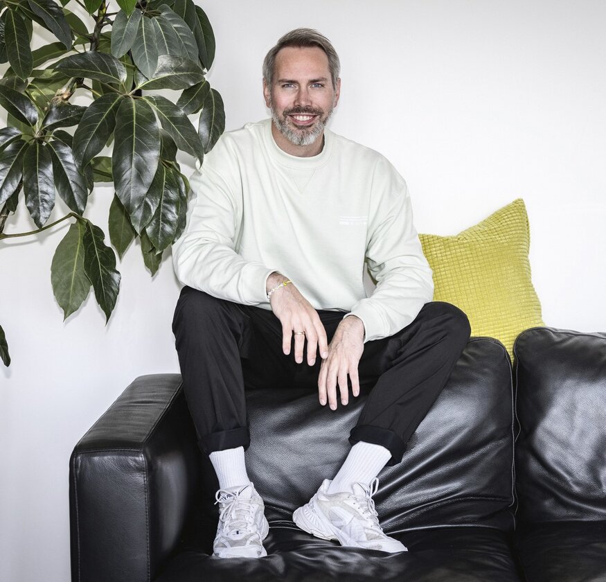 Ein Mann mit grauem Bart sitzt auf der Rückenlehne eines schwarzen Glattledersofas. Rechts neben ihm steht eine große grüne Topfpflanze, links von ihm liegt ein gelbes Sofakissen.