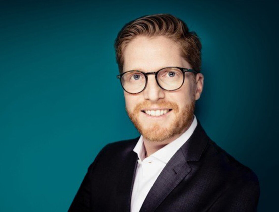 Ein rothaariger Mann mit Bart lächelt in die Kamera. Er trägt schwarze Hornbrille, ein weißes Hemd, darauf ein blaues Sakko. 