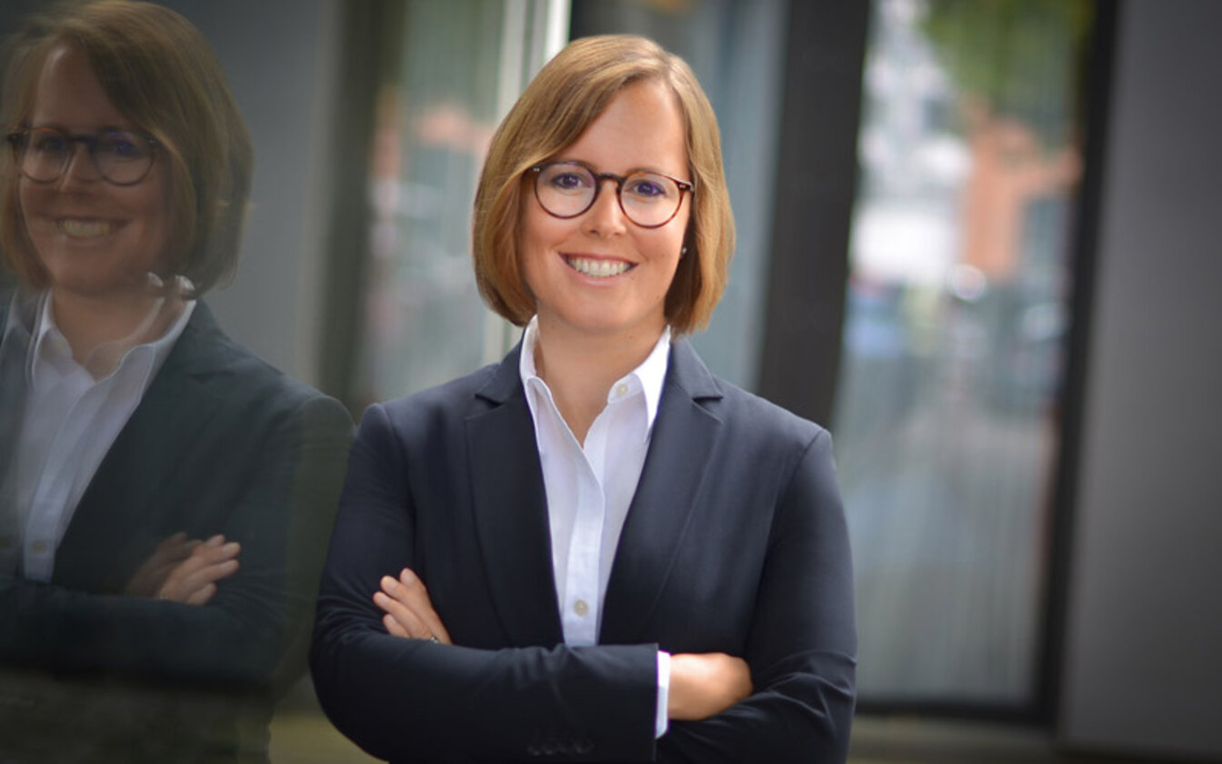 Miriam Fege lehnt lächelnd mit der rechten Schulter an einem Fenster. Die Rothaarige hat einen Bob und trägt eine braun-schwarz-melierte Hornbrille. Sie ist mit einer weißen bluse und einem blauen Blazer gekleidet. Ihre Arme sind verschränkt. 
